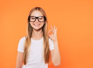 Jeune fille qui sourit avec ses lunettes sur le nez, devant un fond orange